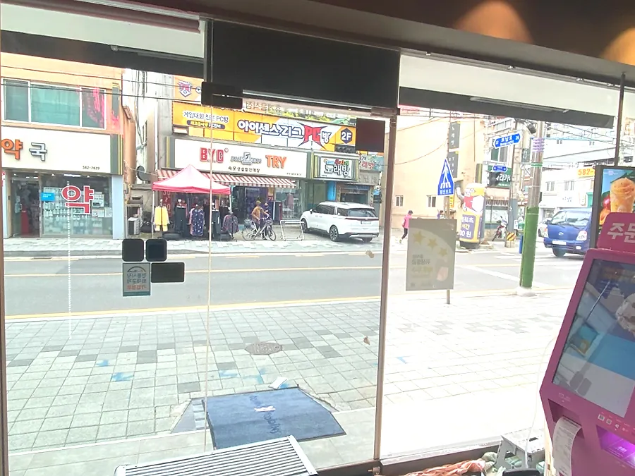 매장 유리문 옆 자외선차단필름 시공 후 도로변 전경과 주문 키오스크가 함께 보이는 모습